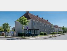 Mehrfamilienhaus in einfacher Bauweise mit grauer Fassade und rotem Satteldach, gelegen an einer ruhigen Straße mit kleinen neu gepflanzten Bäumen und einem Müllsammelplatz im Vordergrund. Die Umgebung ist von gepflegten Grünflächen und vereinzelten weiteren Gebäuden geprägt.