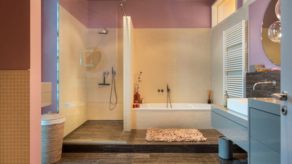 Modern bathroom with purple upper walls and small beige mosaic tiles, featuring a glass walk-in shower, built-in bathtub, and dark tile flooring. A floating vanity with rectangular sink, round mirror, and white towel radiator sit beside the tub, with small rugs on a raised platform.