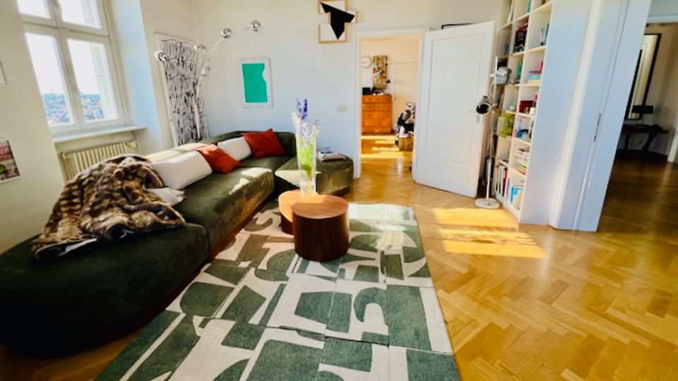 Helles Wohnzimmer mit großen Fenstern, dunklem Ecksofa und einem runden Couchtisch auf einem gemusterten Teppich. Rechts stehen hohe, eingebaute Bücherregale neben einer offenen Tür, und der Raum hat Parkettboden und helle Wände.