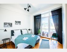 Helles Schlafzimmer mit einem Doppelbett, dekoriert mit weißen Kissen und einer türkisfarbenen Decke, daneben moderne Nachttische und Lampen. Große Fenster mit schwarzen Vorhängen führen auf einen Balkon mit Blick auf ein Gebäude und fließendes Wasser.