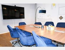Konferenzraum mit einem rechteckigen Holztisch, umgeben von Drehstühlen mit blauen Polstern. An der Wand befindet sich ein großer Bildschirm, daneben eine weiße Wand mit minimalistischen Illustrationen und ein Stehlampe mit blauem Lampenschirm.