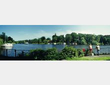 Ein Flussufer mit üppiger Vegetation im Vordergrund, einer Metallbrüstung und zwei Personen, von denen eine angelt. Im Hintergrund sind Boote auf dem Wasser zu sehen, umgeben von Bäumen und kleinen Gebäuden entlang des Ufers. Der Himmel ist blau und klar.