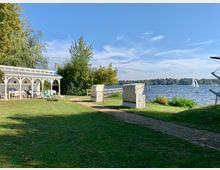 Grüne Uferlandschaft mit einer Rasenfläche, auf der ein kleiner Pavillon mit weißen Holzstühlen und Sonnenliegen steht. Im Hintergrund sind ein See mit einem Segelboot sowie weitere Bäume am gegenüberliegenden Ufer zu sehen.