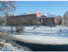 Backsteingebäude in einer verschneiten Winterlandschaft, umgeben von kahlen Bäumen und einem teils zugefrorenen Fluss im Vordergrund. Der Himmel ist klar und blau.