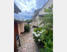 Ein gepflasterter Gartenweg führt an einem cremefarbenen Haus mit dunklem Dach vorbei. Auf der rechten Seite sind blühende Sträucher und grüne Pflanzen, während links eine Garage oder ein Nebengebäude sowie ein Grill sichtbar sind. Ein bewölkter Himmel befindet sich im Hintergrund.