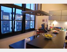 Moderne Küche mit einer großen Arbeitsfläche aus dunklem Material, einem Dunstabzug über der Kochinsel und verschiedenen Küchenutensilien. Im Hintergrund sind große Fenster mit Blick auf ein modernes Wohngebäude zu sehen.