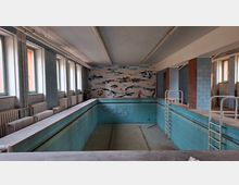 Leeres, gefliestes Hallenbad mit rechteckigem Becken, Metallleitern und umlaufendem Beckenrand. Links stehen hohe Fenster über Heizkörpern, an der Stirnwand ist ein Mosaikbild, und sichtbare Rohrleitungen verlaufen unter der Decke.