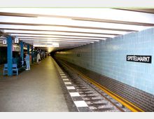 U-Bahn-Station mit einem langen, schmalen Bahnsteig, umgeben von hellblau gefliesten Wänden. Das Schild "Spittelmarkt" ist an der rechten Wand angebracht, Gleise verlaufen mittig, und Passagiere warten an der linken Seite.