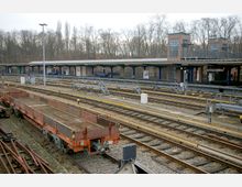 Bahnhof mit mehreren Gleisen und abgestellten Güterwaggons im Vordergrund. Im Hintergrund befinden sich überdachte Bahnsteige mit roten Backsteingebäuden und kahlen Bäumen.