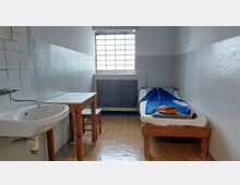 Schlichte Zelle mit einem Einzelbett, einem kleinen Holztisch, einem Hocker und einem Waschbecken an der linken Wand. Ein vergittertes Fenster an der hinteren Wand lässt gedämpftes Tageslicht in den Raum.