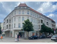 Eckgebäude im Stil der Gründerzeit mit verzierten Fassaden, roten Ziegeldächern und integrierten Gewerbeflächen im Erdgeschoss, darunter ein Fielmann-Geschäft. Davor Gehweg, mehrere Bäume, parkende Autos und Fußgänger.