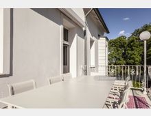 Helle Terrasse mit einem langen weißen Tisch und Stühlen, angrenzend an eine weiße Gebäudewand mit Fenstern und einer Tür; im Hintergrund ein schmiedeeisernes Geländer und Bäume.