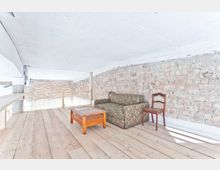 Dachgeschossraum mit sichtbaren Ziegelwänden und heller Holzplankenboden. Im Mittelpunkt stehen ein grünes, gemustertes Sofa, ein Holztisch und ein einzelner Holzstuhl; der Raum wirkt minimalistisch und lichtdurchflutet.