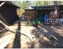 Außenbereich mit einem kleinen Holzgebäude auf der linken Seite und einem überdachten Lagerplatz rechts, der Brennholz, Werkzeuge und Leitern beherbergt. Im Vordergrund befinden sich Gartenschläuche, eine Schubkarre und grüne Gießkannen, umgeben von sandigem Boden und Bäumen im Hintergrund.