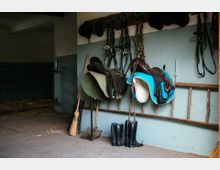 Ein Pferdestall mit Sätteln auf einer Wandhalterung, Zaumzeug, Reitstiefeln und Besen auf dem Boden. Im Hintergrund steht eine Metallbox mit Stroh.