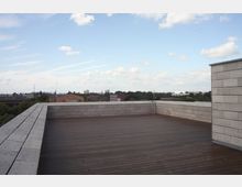 Eine Dachterrasse mit Holzbelag und einer niedrigen Brüstung aus grauen Betonplatten bietet Ausblick auf eine Stadtlandschaft mit vereinzelten Bäumen, Gebäuden und einem teilweise bewölkten Himmel.