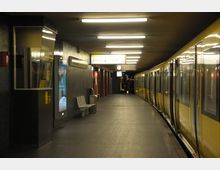U-Bahn-Station mit einem gelben Zug, der an einem leicht gebogenen Bahnsteig hält. Der Bahnsteig ist beleuchtet und mit Sitzbänken, Wandanzeigen, Fahrplänen und einer analogen Uhr ausgestattet.