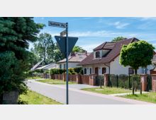 Straßenszene in einem Wohngebiet mit Einfamilienhäusern, Zäunen und Vorgärten. Im Vordergrund steht ein Straßenschild mit den Namen "Havelländer Weg" und "Grüner Straße", im Hintergrund sind Bäume und ein klarer Himmel zu sehen.