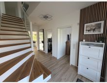 Moderner Eingangsbereich eines Hauses mit einer Holztreppe auf der linken Seite, hellem Laminatboden und weißen Wänden. Rechts steht ein weißes Schuhregal, darüber eine Holzlamellenwand mit einem Bild von Wildtieren. Im Hintergrund sind offene Türen zu anderen Räumen erkennbar.