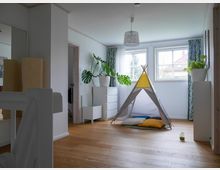 Heller Raum mit einem Holzfußboden, einem kleinen Kinderspielzelt in der Mitte und bunten Kissen darin. Im Hintergrund stehen weiße Kommoden, Zimmerpflanzen und es gibt zwei große Fenster mit Blick nach draußen.