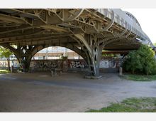 Ein metallisches Brückengerüst mit geschwungenen Trägern überspannt eine sandige, parkähnliche Fläche. Unter der Brücke sind Wände mit Graffitis bedeckt, und im Hintergrund sind Gleisanlagen sowie ein Bahnhofsgebäude sichtbar.