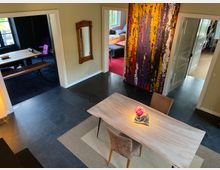 Esszimmer in einem modernen Wohnhaus mit dunklen Fliesenboden, einem Holztisch und zwei Stühlen. Im Hintergrund sind weitere Räume mit buntem Teppich und einem abstrakten, farbenfrohen Gemälde an der Wand sichtbar.