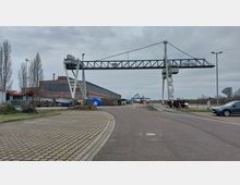 Industriehafen mit einem großen Portalkran, rechts neben einer gepflasterten Straße. Im Hintergrund sind Lagerhallen, gestapelte Materialien und weitere Kräne zu sehen, umgeben von Bäumen und leerer Landschaft.