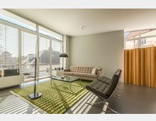 Modernes Wohnzimmer mit einer großen Fensterfront, durch die Tageslicht einfällt, einer beigefarbenen Couch, zwei schwarzen Sesseln und einem grünen Teppich unter einem Glastisch. Im Hintergrund steht ein bogenförmiger Stehlampenbogen, links ein niedriger Sideboard mit einem Radio und rechts ein gelber Vorhang.