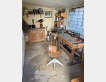 Werkstattraum mit rustikalem Charme: ein hölzerner Arbeitstisch mit Werkzeugen, ein alter Drehstuhl, Regale mit Metall- und Holzwaren sowie eine Glasbausteinfensterwand, die natürliches Licht hereinlässt. Der Boden besteht aus unregelmäßig verlegten Steinfliesen.