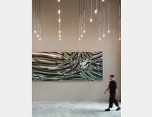 Minimalistisch gestalteter Innenraum mit einer abstrakten Wandkunst aus mehreren Paneelen mit wellenförmigen, metallisch schimmernden Mustern. Von der Decke hängen zahlreiche schlanke Pendelleuchten mit warmem Licht, während eine Person in schwarzer Kleidung durch den Raum läuft.