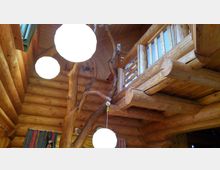 Innenraum einer Blockhütte mit sichtbaren Holzwänden und -balken. Im Vordergrund hängen runde, leuchtende Lampen, und eine Baumstammkonstruktion dient als dekoratives Element und Stütze für eine Holzbalkon-Ebene.