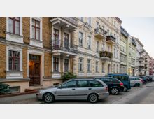 Reihe von mehrstöckigen Altbauwohnhäusern mit verzierten Fassaden, Balkonen und großen Fenstern; im Vordergrund parken mehrere Autos entlang der Straße.
