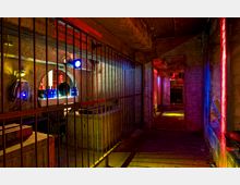 Industrieller Raum mit Betonwänden und Decke, beleuchtet von bunten Lichtquellen in Rot, Blau und Gelb. Links befinden sich Metallgitter, hinter denen eine Anrichte mit Flaschen sowie runde Öffnungen in der Wand sichtbar sind; im Hintergrund setzen sich Gänge und weitere Gitter fort.