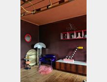 Wohnzimmer im Retro-Stil mit dunklen Wänden, einem blauen Designerstuhl, einer weißen Lampe, einer flauschigen pinken Bodenmatte und Möbeln aus Holz. Im Raum befinden sich skulpturale Dekorobjekte auf Regalen und ein Holzlattenelement an der Decke.