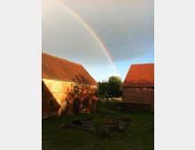 Ein ländlicher Hof mit zwei Backsteingebäuden und roten Ziegeldächern, dazwischen ein grüner Hofbereich mit einer Sitzbank und dekorativer Vegetation. Im Hintergrund ist ein Regenbogen vor bewölktem Himmel zu sehen.