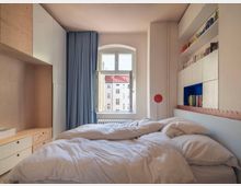 Ein helles Schlafzimmer mit Doppelbett, weißen Bettdecken und einem großen Fenster mit Blick auf ein mehrstöckiges Wohngebäude. Links befinden sich Holzschränke und rechts eine eingebaute Regalwand mit Büchern, teilweise in weißen Schränken integriert.