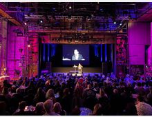 Ein Veranstaltungsraum mit einer Bühne, auf der eine Person spricht, während sie auf einer Leinwand im Hintergrund übertragen wird. Der Raum ist mit Industrieelementen wie Metallgerüsten und sichtbaren Rohren gestaltet, beleuchtet in violetten und blauen Farbtönen, mit einem Publikum, das an Tischen sitzt.