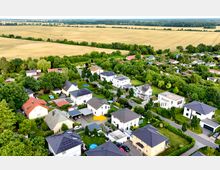 Luftaufnahme eines Wohngebiets mit Einfamilienhäusern, umgeben von Gärten und Grünflächen. Im Hintergrund erstrecken sich weite Felder und eine Baumreihe.