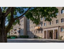 Wohngebäude mit mehreren Stockwerken und Balkonen, umgeben von gepflastertem Gehweg und grüner Bepflanzung; im Vordergrund ein großer Baum, der Schatten wirft. Ein Durchgangsportal aus Stein prägt den Eingangsbereich des Gebäudes.