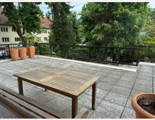 Freiluft-Terrasse mit grauem Steinboden, einem Holztisch und Blumentöpfen. Im Hintergrund sind grüne Bäume, gelb verputzte Gebäude mit roten Dächern und eine metallene Geländer-Umrandung zu sehen.