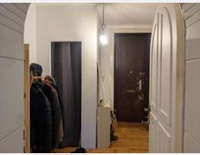 Flur einer Wohnung mit Holzboden und einer dunkelbraunen Eingangstür. Links steht eine Kleiderstange mit Jacken und Mänteln, rechts ein weißer geöffneter Türrahmen; eine einzelne Glühbirne hängt von der Decke.