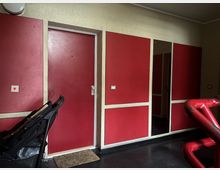 Ein kleiner Innenraum mit roten Wänden und weißen Rahmen, einer Tür in der Mitte und einem Kinderwagen im Vordergrund links. Rechts im Bild ist ein rotes Geländer sichtbar, und der Boden besteht aus dunklem Material mit leicht reflektierender Oberfläche.