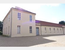 Zweistöckiges, beige gestrichenes Gebäude mit Satteldach und weißen Fenstern, daneben ein eingeschossiger Flügel mit roten Ziegeln. Der Vorplatz ist ein großer, offener Kiesbereich, und im Hintergrund sind vereinzelt Bäume sichtbar.