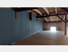 Ein leerer Dachbodenraum mit einer schrägen Holzbalkendecke, einer dunkel gestrichenen Wand auf der linken Seite und einer Ziegelwand mit einem großen, beleuchteten Fenster auf der Rückseite. Der Boden ist mit Holzplatten bedeckt, und freiliegende Holzbalken stützen die Konstruktion.