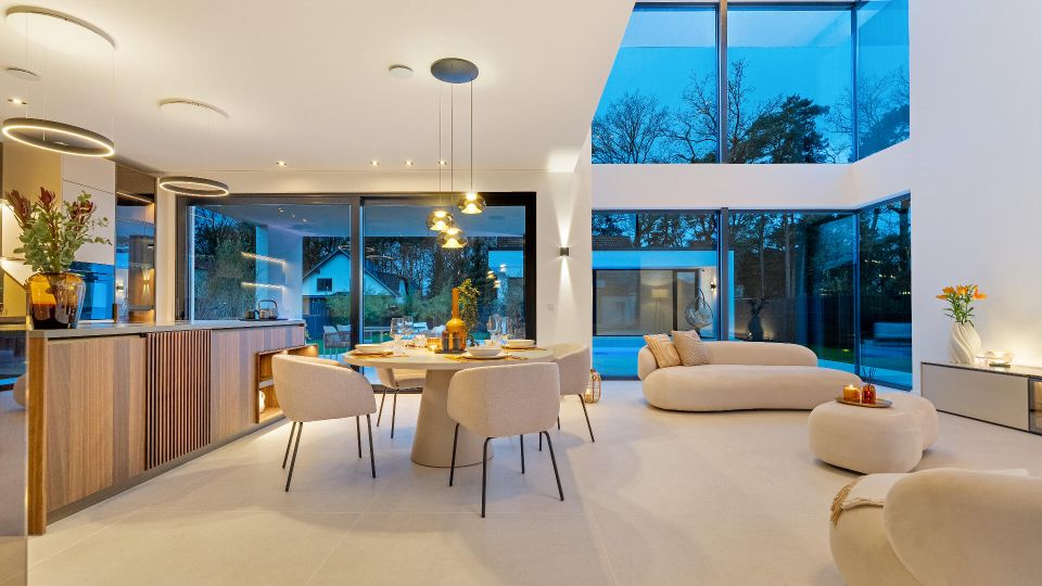 Offener, moderner Wohn- und Essbereich mit integrierter Küche, hellen Fliesen und minimalistischem Mobiliar, darunter runder Esstisch und gepolsterte Sitzmöbel. Große bodentiefe Glasfronten und ein zweigeschossiger Fensterbereich geben den Blick in den Garten frei; mehrere Pendelleuchten und indirekte Deckenbeleuchtung strukturieren den Raum.