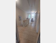 Ein heller Flur in einem Wohnhaus mit cremefarbenen Fliesen und hellen Wänden. Rechts ist eine Tür, links ein Geländer, und am Ende des Flurs befinden sich zwei Glastüren mit Blick ins Freie. Ein kleines Zelt steht auf dem Boden nahe den Türen.