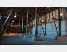 Ein großer, offener Raum in einer historischen Halle mit sichtbaren Holzstützen, Backsteinwänden und einem Holzgebälk im Dach. Mehrere Hängeleuchten hängen von der Decke, und der Boden besteht aus Beton.