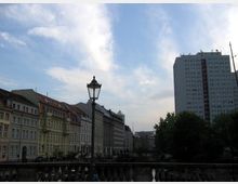 Straßenansicht mit historischen mehrstöckigen Gebäuden auf der linken Seite und einem modernen Hochhaus auf der rechten, im Vordergrund ein Laternenmast und ein Geländer; im Hintergrund ein Kanal und Bäume unter einem leicht bewölkten Himmel.