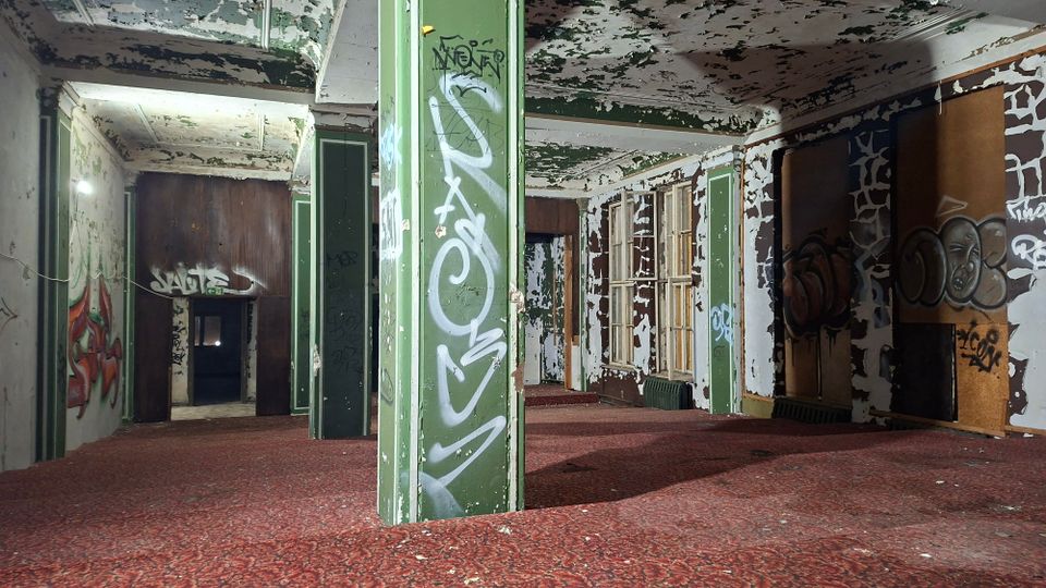 Innenraum eines verlassenen Gebäudes mit abblätternder Farbe an Decke und Wänden, graffitibeschmierten grünen Stützpfeilern und rotem Teppichboden. An der rechten Seite stehen Fensterreihen und Heizkörper, im Hintergrund führen offene Durchgänge in weitere dunkle Räume.