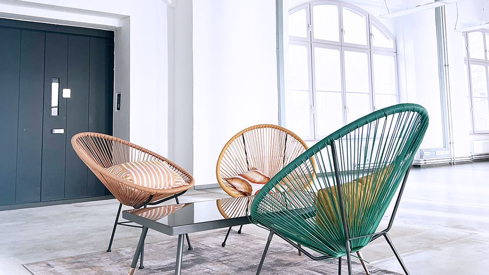 Ein moderner Innenraum mit hohen Decken, weißen Wänden und großen Rundbogenfenstern. Im Vordergrund stehen drei bunte Stühle im Retro-Stil um einen kleinen Glastisch auf einem gemusterten Teppich.